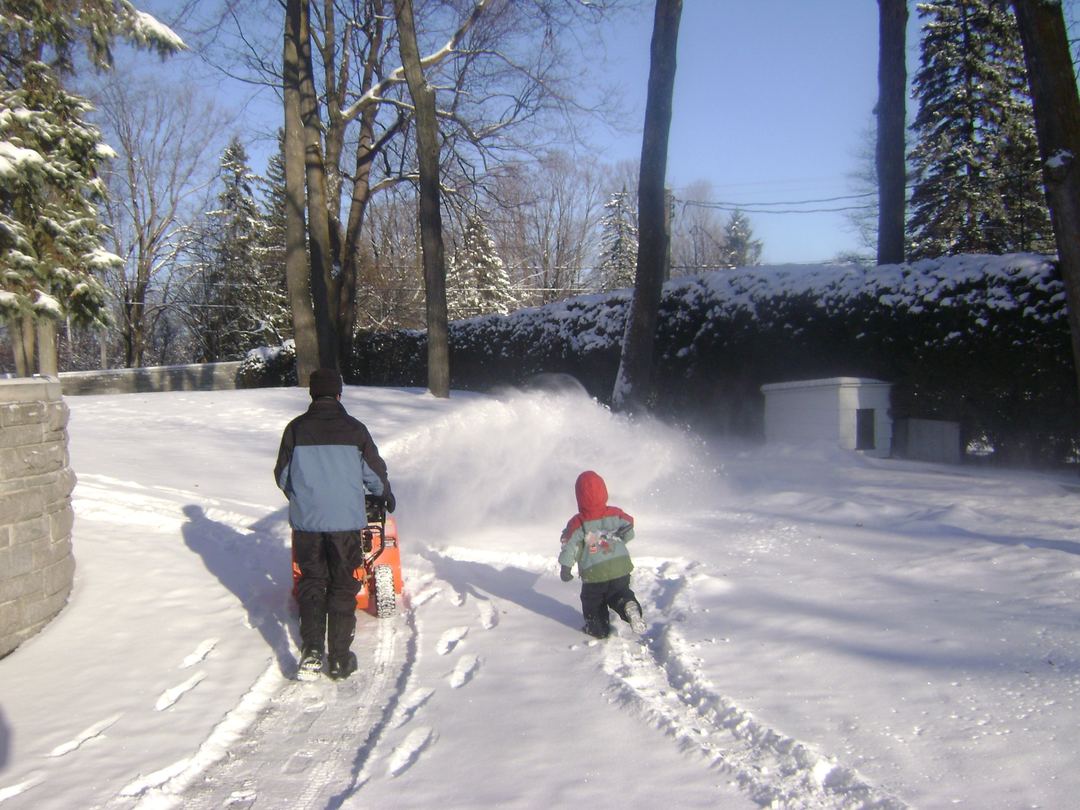 Un jeune enfant au capuchon rouge suit son père dans la neige, sur le chemin que celui-ci vient de dégager.